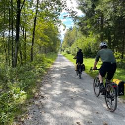 Zwei Radfahrer mit Packtaschen fahren auf einem Schotterweg durch einen grünen Wald in Böhmen, Tschechien.