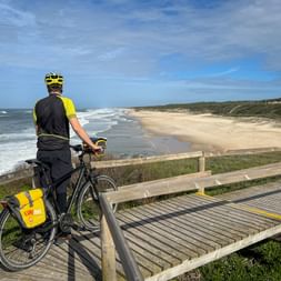 Cyclist with yellow panniers on wooden boardwalk overlooking sandy beach and Atlantic Ocean on Northern Portugal's coastal route.