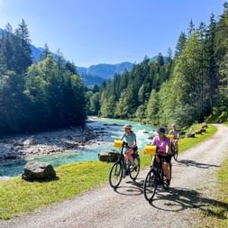 Three cyclists with yellow panniers on a path beside the turquoise Saalach River near Lofer, surrounded by dense forest and mountains.