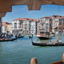 Blick unter einer Brücke auf Gondeln und Boote auf dem Canal Grande in Venedig, gesäumt von historischen bunten Gebäuden unter blauem Himmel.