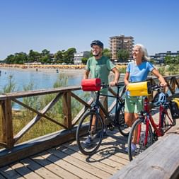 Two cyclists with bikes on wooden bridge overlooking beach in Grado. Sandy beach with sunbathers and buildings visible in background under blue sky.
