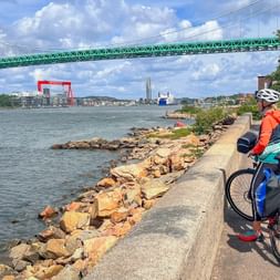 Cyclist with helmet and panniers on waterfront path in Gothenburg, viewing harbor with green suspension bridge and red crane under blue sky.