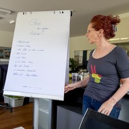 Red-haired woman in gray shirt standing beside flipchart with 'Ghee' written at top and handwritten notes in German in modern kitchen.