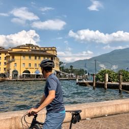 Cyclist with helmet riding along the waterfront promenade in Riva del Garda with yellow historic buildings, harbor, and mountains in background.