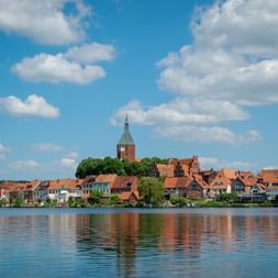 View across the lake to Mölln's historic town center with red-tiled roofs, a prominent church tower, and green trees under a blue sky.
