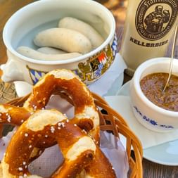 Traditional Bavarian breakfast with white sausages in a ceramic bowl, soft pretzels with salt, sweet mustard, and Kellerbier on a wooden table.