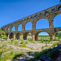 The Pont du Gard, a three-tiered Roman aqueduct bridge with golden stone arches spanning a dry riverbed in Provence under a clear blue sky.