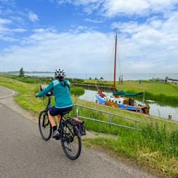 Radfahrerin in türkisfarbener Jacke fährt auf Deichweg neben Kanal mit traditionellem Segelboot im grünen Schilf unter blauem Himmel.