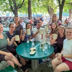 Group of nine women from Eurohike team sitting around green table in beer garden, raising beer mugs in celebration under leafy trees.