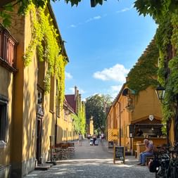 Narrow cobblestone street in Fuggerei, Augsburg, lined with yellow row houses covered in green ivy. Cyclists and pedestrians visible under blue sky.