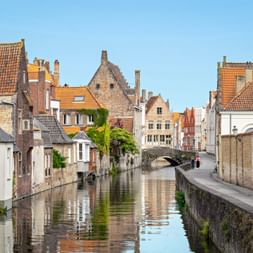 Scenic canal in Bruges with medieval brick buildings, orange-tiled roofs, and a stone bridge. Calm water reflects the architecture under clear sky.