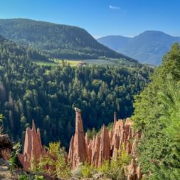 Erdpyramiden mit Steinkappe am Ritten, Südtirol, umgeben von Wald mit Bergpanorama und blauem Himmel im Hintergrund.