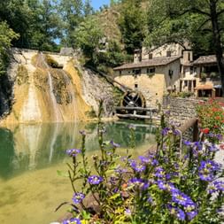 Wassermühle Molinetto della Croda mit Wasserrad neben einem Teich mit Wasserfall. Lila und rote Blumen blühen entlang des Kieswegs, umgeben von Bäumen.