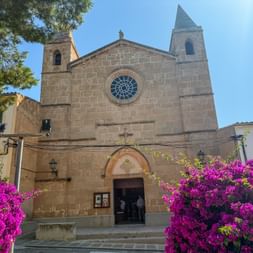 Stone church with round rose window and bell tower in Porto Colom, Mallorca, framed by vibrant pink bougainvillea flowers under blue sky.