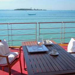 Outdoor terrace on MS Ave Maria with wooden table, chairs with white cushions, and coffee service overlooking the turquoise Venetian lagoon.