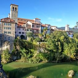 Historic buildings of Cividale del Friuli perched on a cliff above a green river, with a stone bridge and church tower visible under blue sky.