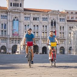 Two cyclists riding across the main square in Trieste with historic white buildings and fountain in the background under clear sky.
