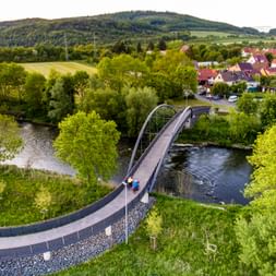 Aerial view of a curved bicycle bridge with metal arch spanning the Werra River, surrounded by green trees and a village with hills in background.