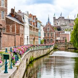 Kanal in Gent mit bunten Blumenkörben entlang der Steinmauern. Historische Backsteingebäude und eine mittelalterliche Burg im Hintergrund.