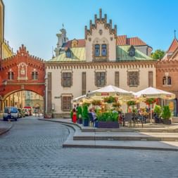 Cobblestone square in Krakow with historic Collegium Maius building, red brick archway, and outdoor café with white umbrellas under blue sky.