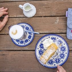 Apple strudel with powdered sugar and whipped cream on a blue-white plate next to a cappuccino and Eurobike pouch on a wooden table.