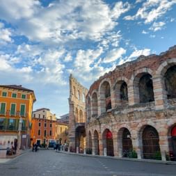 Antikes römisches Amphitheater Arena von Verona mit rosa-weißen Steinbögen. Bunte Gebäude säumen den Kopfsteinpflasterplatz unter blauem Himmel mit Wolken.