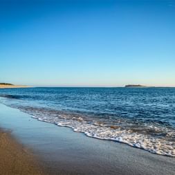 Sandy beach in Caminha with gentle waves and blue water. Forte da Ínsua fortress visible on small island in the distance under clear sky.