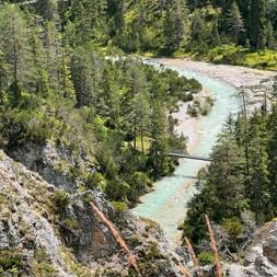 Türkisfarbene Isar fließt durch steiniges Flussbett, umgeben von dichtem grünen Wald. Eine kleine Fußgängerbrücke überquert den Gebirgsbach.