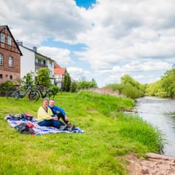 Zwei Radfahrer sitzen auf einer Decke am Flussufer bei Lobenhausen. Fahrräder auf Grasfläche, Fachwerkhäuser im Hintergrund unter bewölktem Himmel.