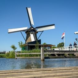 Traditional Dutch windmill with white sails at Kinderdijk beside a canal with wooden bridge and Dutch flag under clear blue sky.