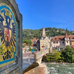 Bridge in Meran with mosaic eagle emblem, crossing green river. Historic town with church tower and hillside buildings under blue sky.