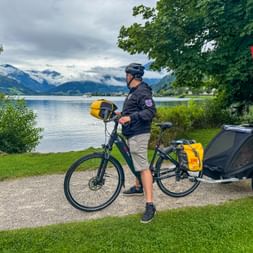 Radfahrer mit Helm steht neben Fahrrad mit gelben Taschen und schwarzem Anhänger am Zeller See. Berge und bewölkter Himmel im Hintergrund.