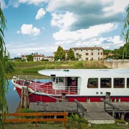 Rot-weißes Passagierschiff Ave Maria am Holzsteg in Portegrandi. Weidenzweige rahmen die Szene mit italienischen Gebäuden im Hintergrund.