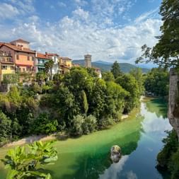 Grüner Fluss durch Cividale del Friuli mit bunten Häusern am Ufer, einem Glockenturm und Bergen im Hintergrund unter blauem Himmel.