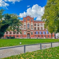 Red brick university building in Rostock with Gothic Revival architecture, surrounded by green lawn and mature trees under blue sky.