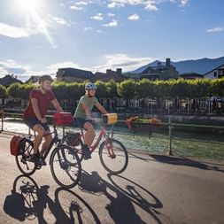 Two cyclists riding along the sunny promenade in Bad Ischl, Salzkammergut. Tree-lined waterfront with flowers and mountains in background.