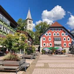 Fußgängerzone in Saalfelden mit bunten Gebäuden, einem roten historischen Haus, Kirchturm und Holzbänken unter blauem Himmel.