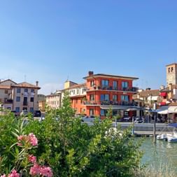 Canal in Marano, Friuli with moored boats, colorful buildings, and a bell tower. Pink flowers bloom in the foreground under a clear blue sky.