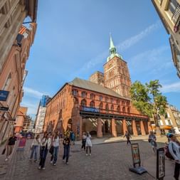 Historic red brick Town Hall in Stralsund with green-topped tower. Pedestrians walk across cobblestone square under blue sky.