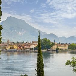 View of Riva del Garda at Lake Garda with colorful buildings along the waterfront, framed by cypress trees, and mountains in the background.