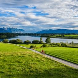 Three cyclists on a path through green meadows near Riegsee lake with mountains in the background under a cloudy sky in Bavaria.