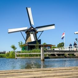 Historische Windmühle mit weißen Flügeln in Kinderdijk neben einem Kanal mit Holzbrücke und niederländischer Flagge unter blauem Himmel.