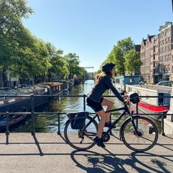 Female cyclist on bridge over Brouwersgracht canal in Amsterdam with traditional Dutch houses, houseboats, and trees lining the waterway.