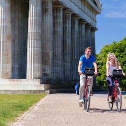 Two cyclists with bikes standing in front of the neoclassical Walhalla temple with its impressive columns in Kehlheim, surrounded by green trees.