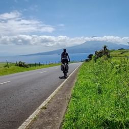 Radfahrer auf Küstenstraße mit grünen Feldern und Blick auf den Vulkan Ponta do Pico über blaues Meer unter teilweise bewölktem Himmel.
