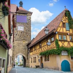 Steinerner Torturm mit Uhr und gelbes Fachwerkhaus an Kopfsteinpflasterstraße in Rothenburg ob der Tauber unter blauem Himmel mit weißen Wolken.