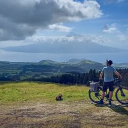 Radfahrer mit E-Bike auf grasbewachsenem Hügel auf den Azoren, Blick auf Vulkanberg über dem Meer unter dramatischem Wolkenhimmel.