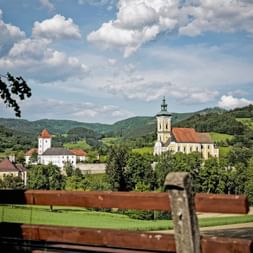Blick auf Strudengau mit gelber Kirche mit Glockenturm und weißem Schloss mit rotem Dach, umgeben von grünen Hügeln und Wäldern unter blauem Himmel.