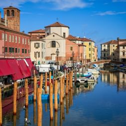 Canale Vena in Chioggia mit bunten Häusern, Kirche, Glockenturm, hölzernen Pfählen und Booten unter blauem Himmel.