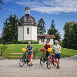 Three cyclists with helmets standing with their bikes in front of the Silent Night Chapel in Oberndorf. The white chapel with dark dome is surrounded by green lawn and trees.